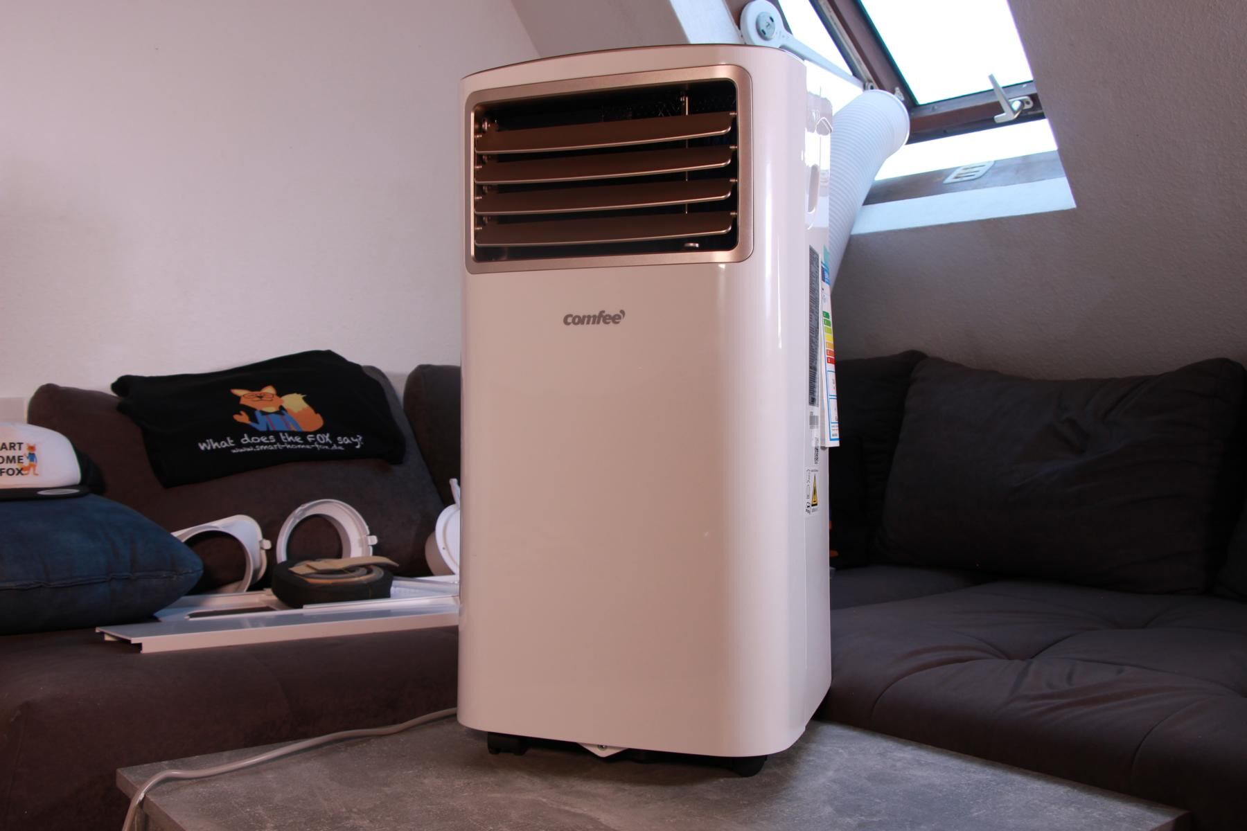 Man sieht ein Monoblock-Gerät von Comfee, der Abluftschlauch wird im Test in einer Mietwohnung durch ein Schrägdachfenster geleitet.